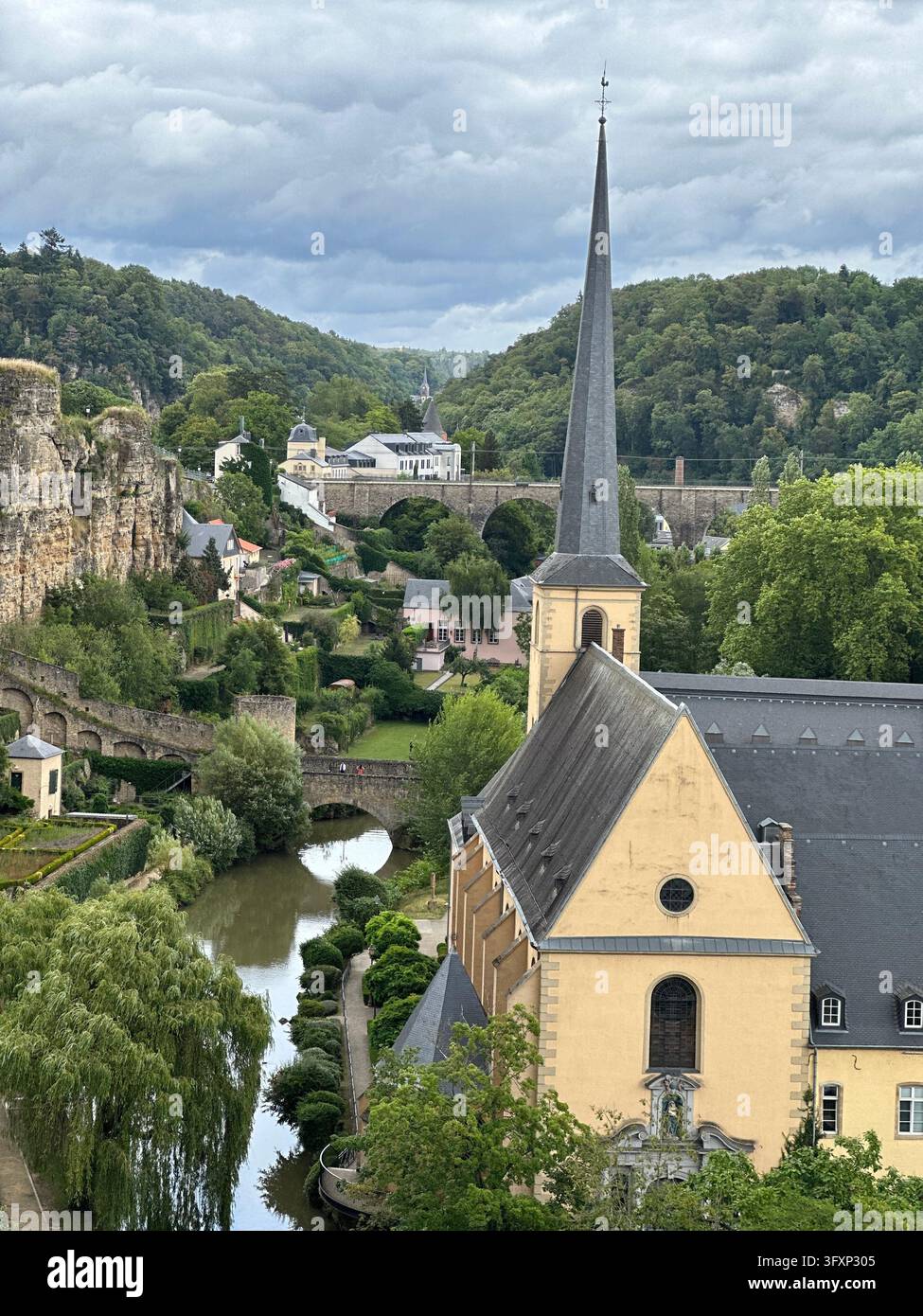 Vista verticale della guglia della chiesa di San Giovanni accanto al fiume e al ponte di pietra nella città di Lussemburgo. L'alta guglia della chiesa si erge sopra una valle fluviale circondata da sto - Immagine stock catturata con smartphone
