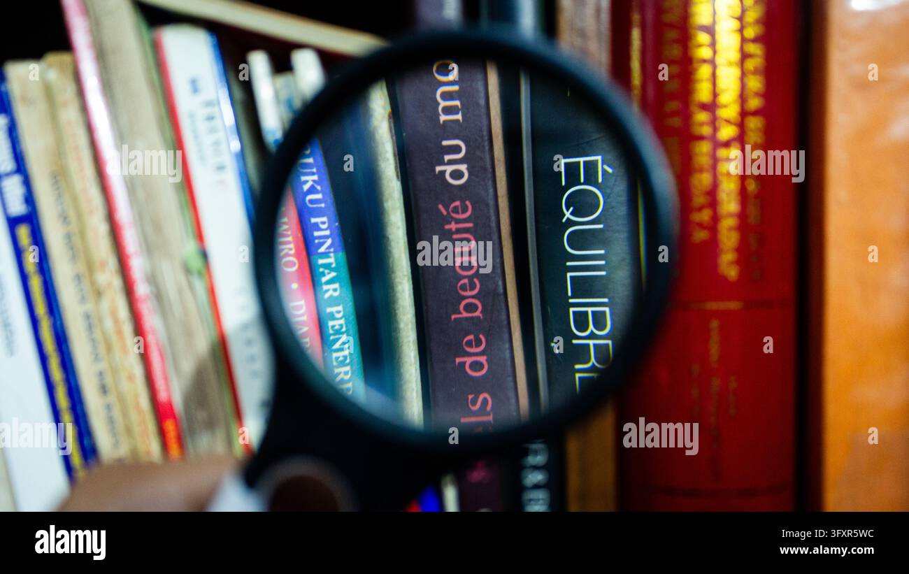Primo piano di una lente d'ingrandimento che esamina i dorsi di libri francesi tra cui “Équilibre” su uno scaffale della biblioteca Foto Stock