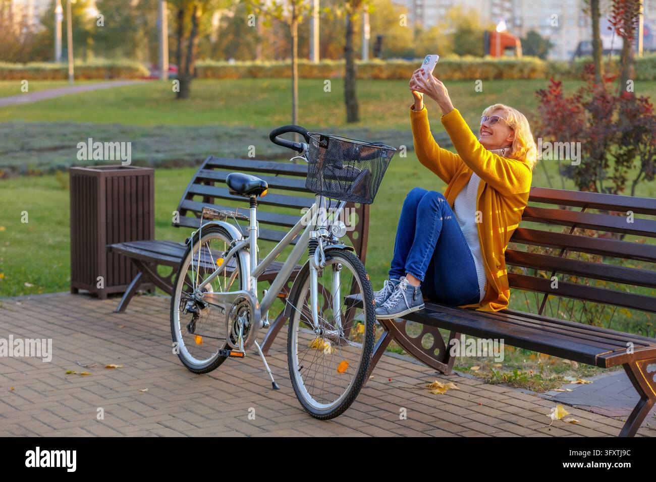 Donna che fa una videochiamata sullo smartphone durante una pausa dal ciclismo nel parco cittadino autunnale, seduta su una panchina accanto alla bicicletta. Comunicazione video, co sociale Foto Stock