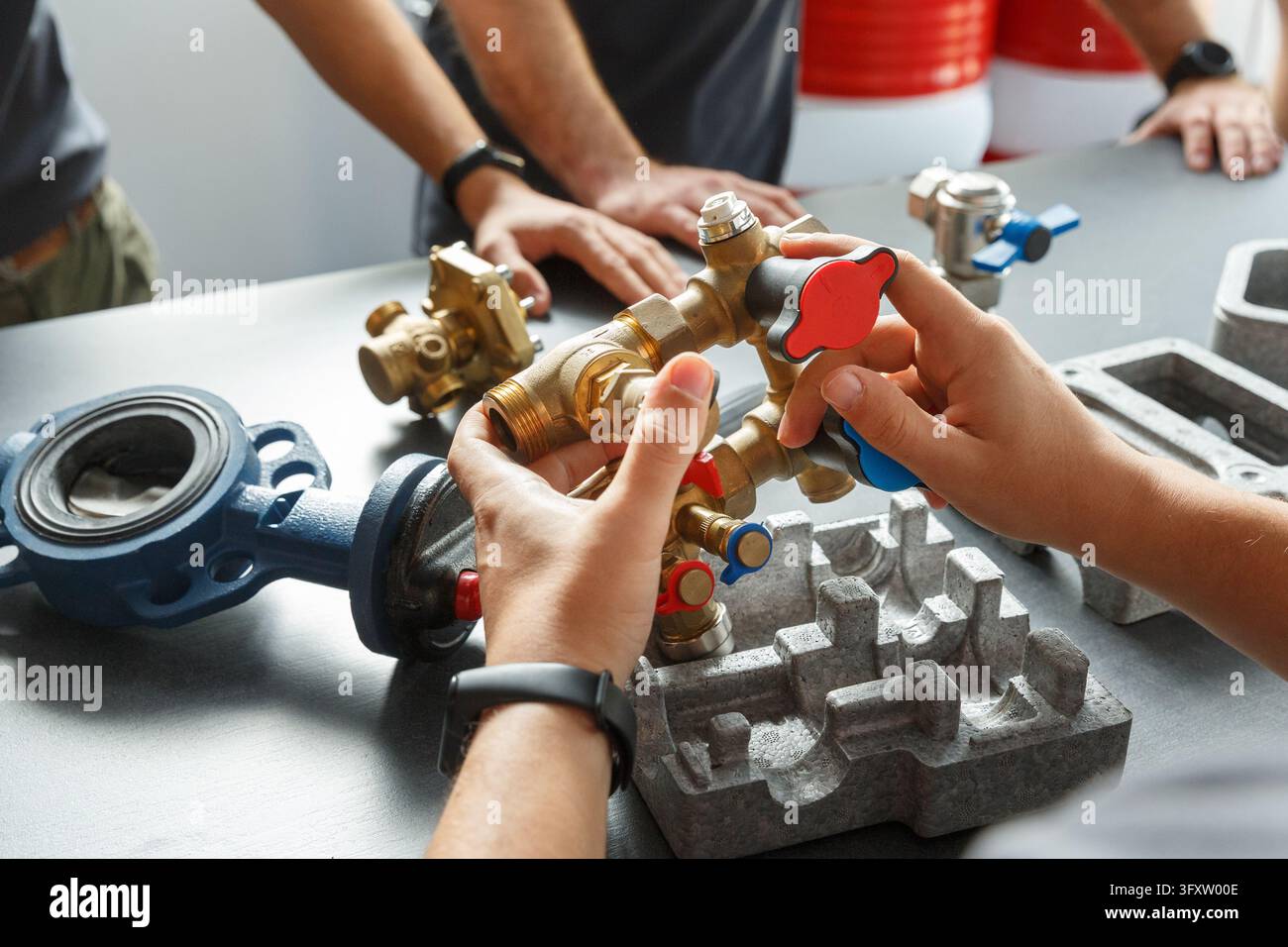 Prima vista di mani che tengono un complesso assemblaggio di valvole in ottone e raccordi di riscaldamento su un tavolo durante un workshop tecnico. Gli ingegneri discutono Foto Stock