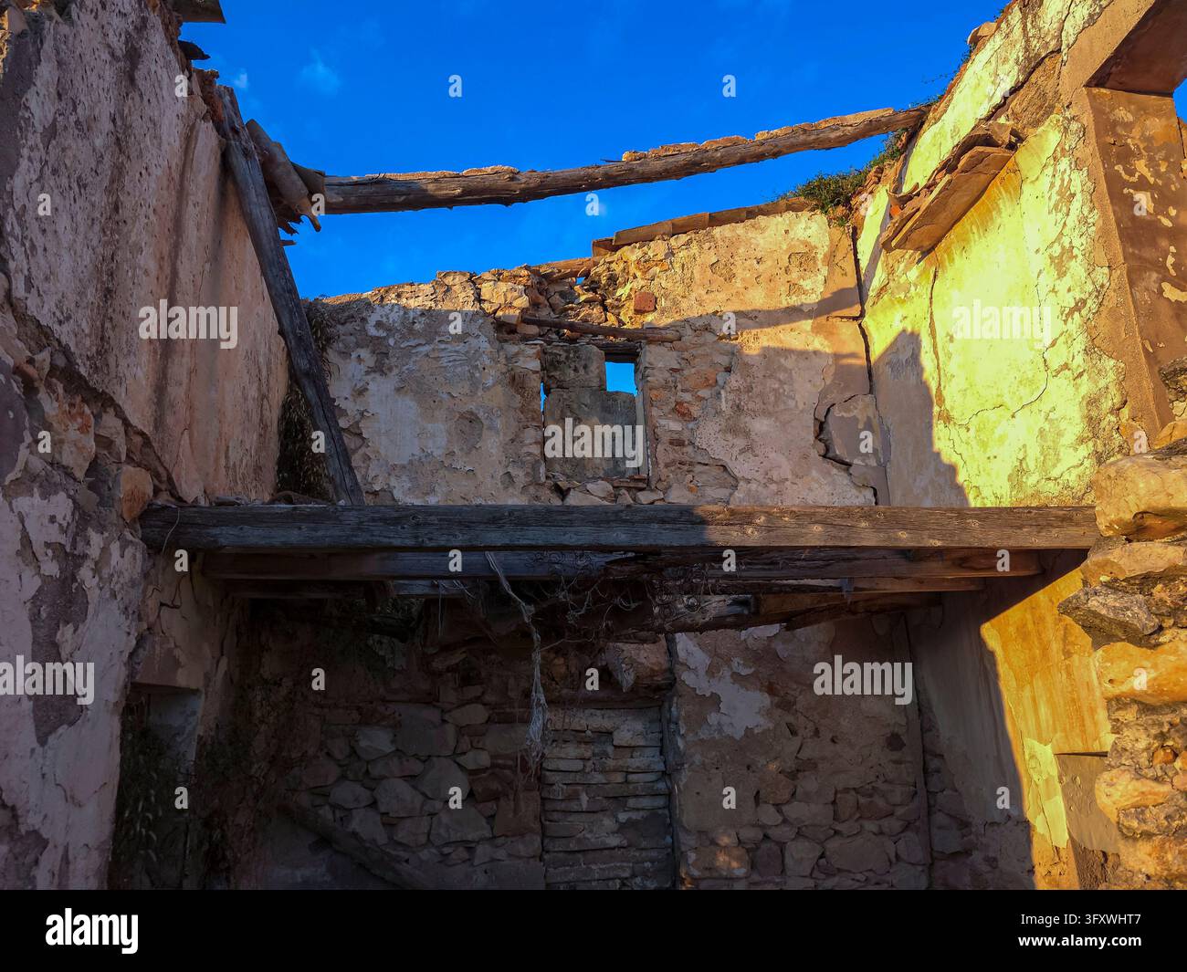 Interno illuminato dal sole di un vecchio edificio di pietra in rovina con travi di legno marcite, muri di intonaco crepati e un tetto crollato che si apre su un cielo azzurro. Foto Stock