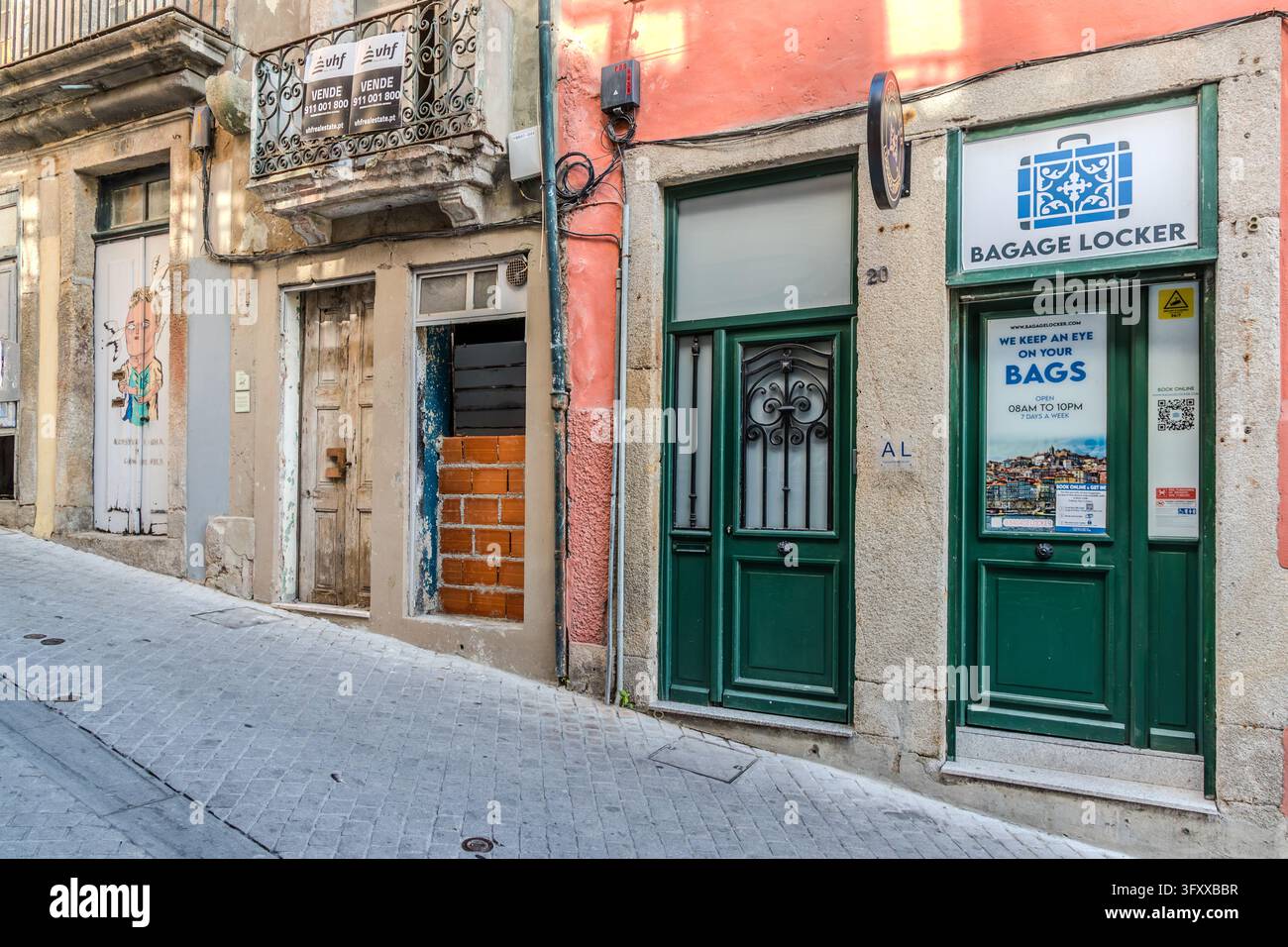 Il negozio “Bagage Locker” si trova in una casa con facciata rosa su una ripida strada acciottolata a Porto, Portogallo. Lì si deposita il bagaglio. Un poster sulla porta verde dice “Teniamo d’occhio le tue borse”. Sotto ci sono gli orari di apertura “Aperto dalle 08:00 alle 22:00, 7 giorni su 7”. A sinistra si trova una casa fatiscente con una finestra murata. Due cartelli sul balcone sopra recitano “Vende”, che significa “In vendita”, e nominano l’agente immobiliare vhf real estate. Foto Stock