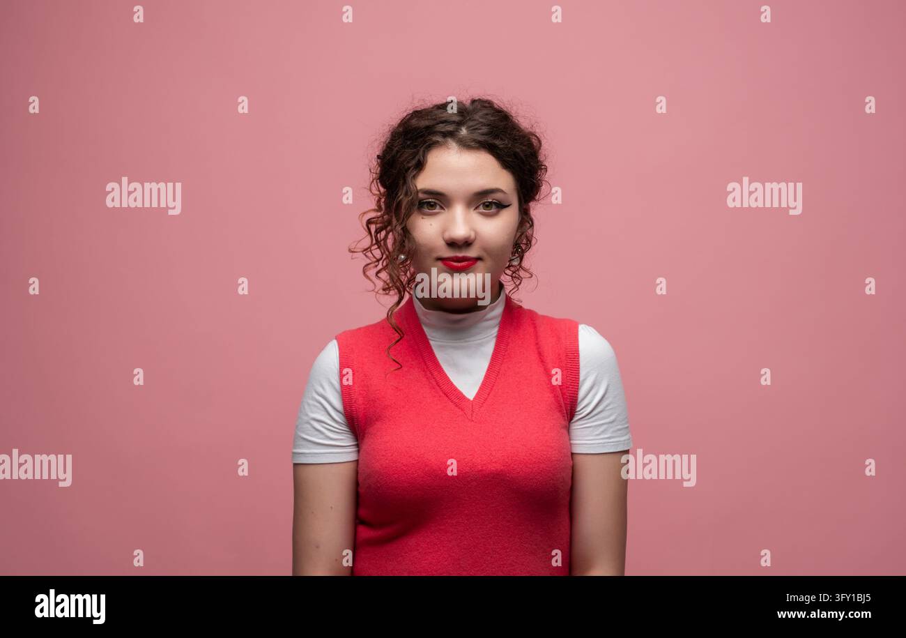 Giovane donna con capelli ricci che indossa un giubbotto rosso e una camicia bianca, posa con sicurezza su uno sfondo rosa Foto Stock