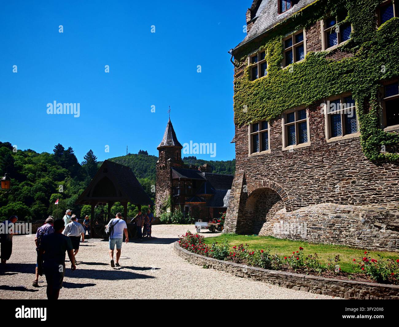 Turisti che camminano attraverso il cortile del castello medievale di Reichsburg Cochem con muri di edera, casa del pozzo e colline verdi in Germania. Foto Stock