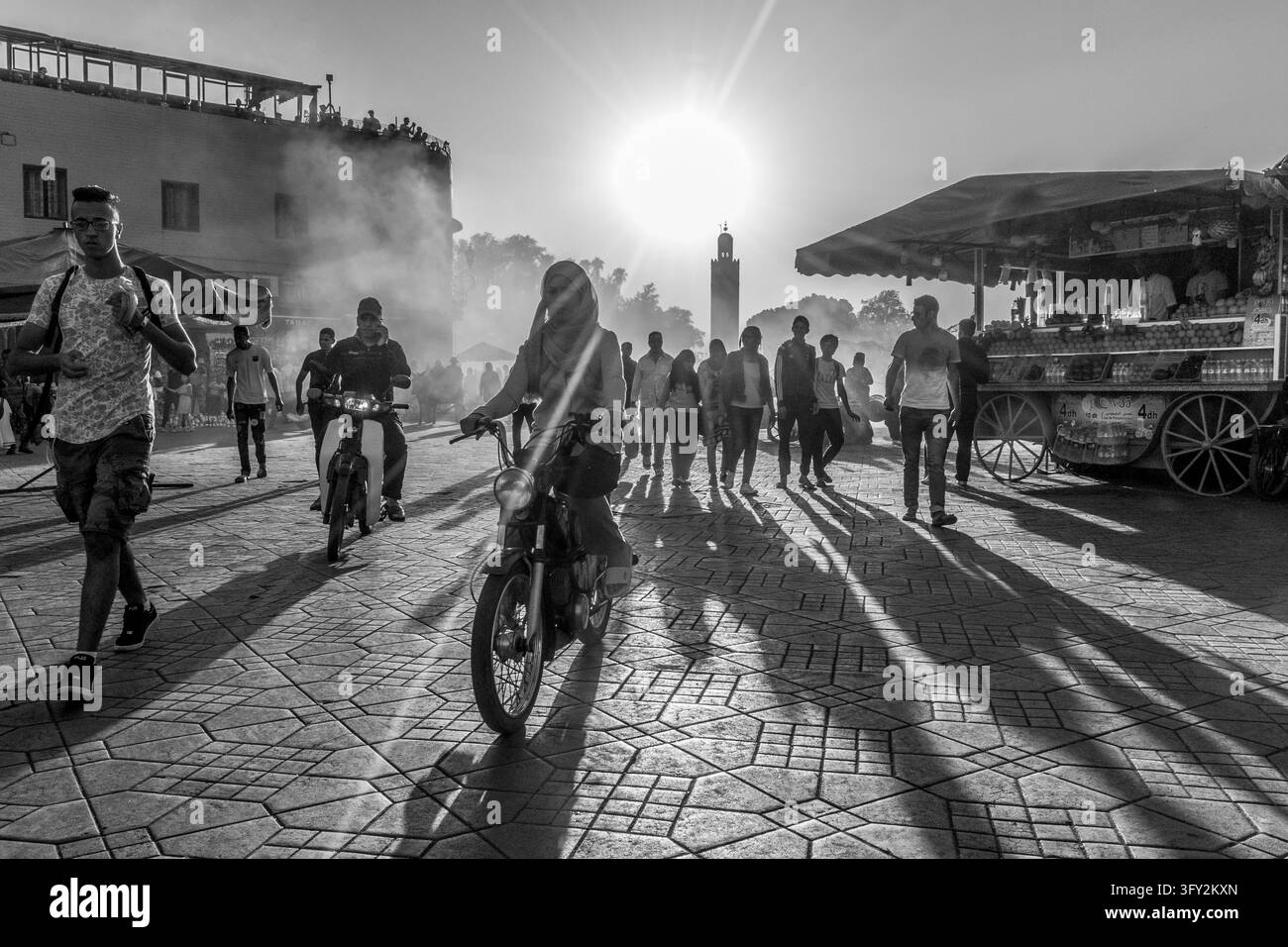 Donna guida un motorino attraverso una piazza affollata di Marrakech mentre i pedoni attraversano la piazza illuminata dal sole vicino alla moschea Koutoubia, illustrando i trasporti urbani, co Foto Stock