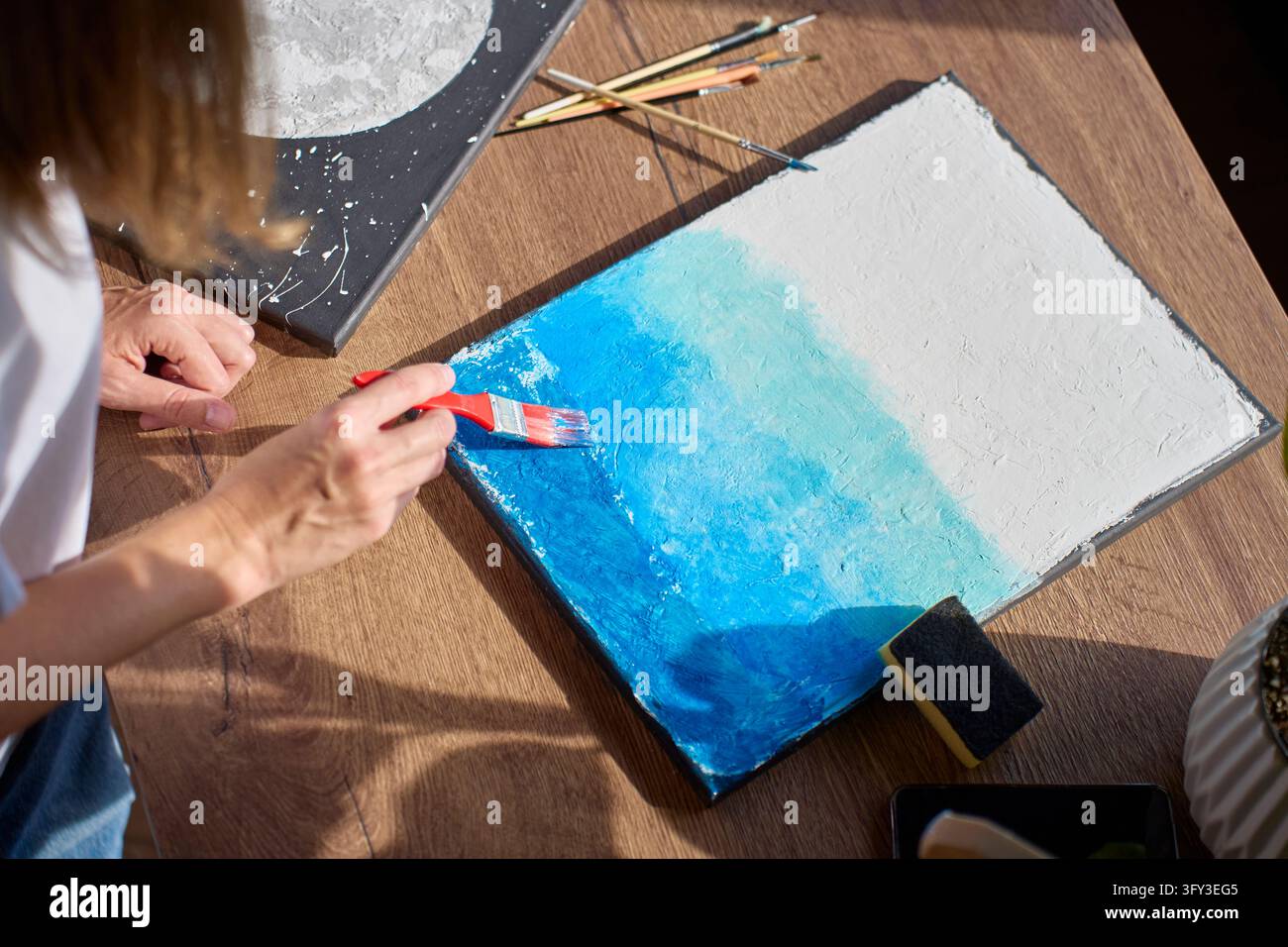 Donna che dipinge diverse sfumature di blu su una tela ruvida. Usa un pennello largo per sfumare i colori. Hobby creativo e creazione di arte Foto Stock