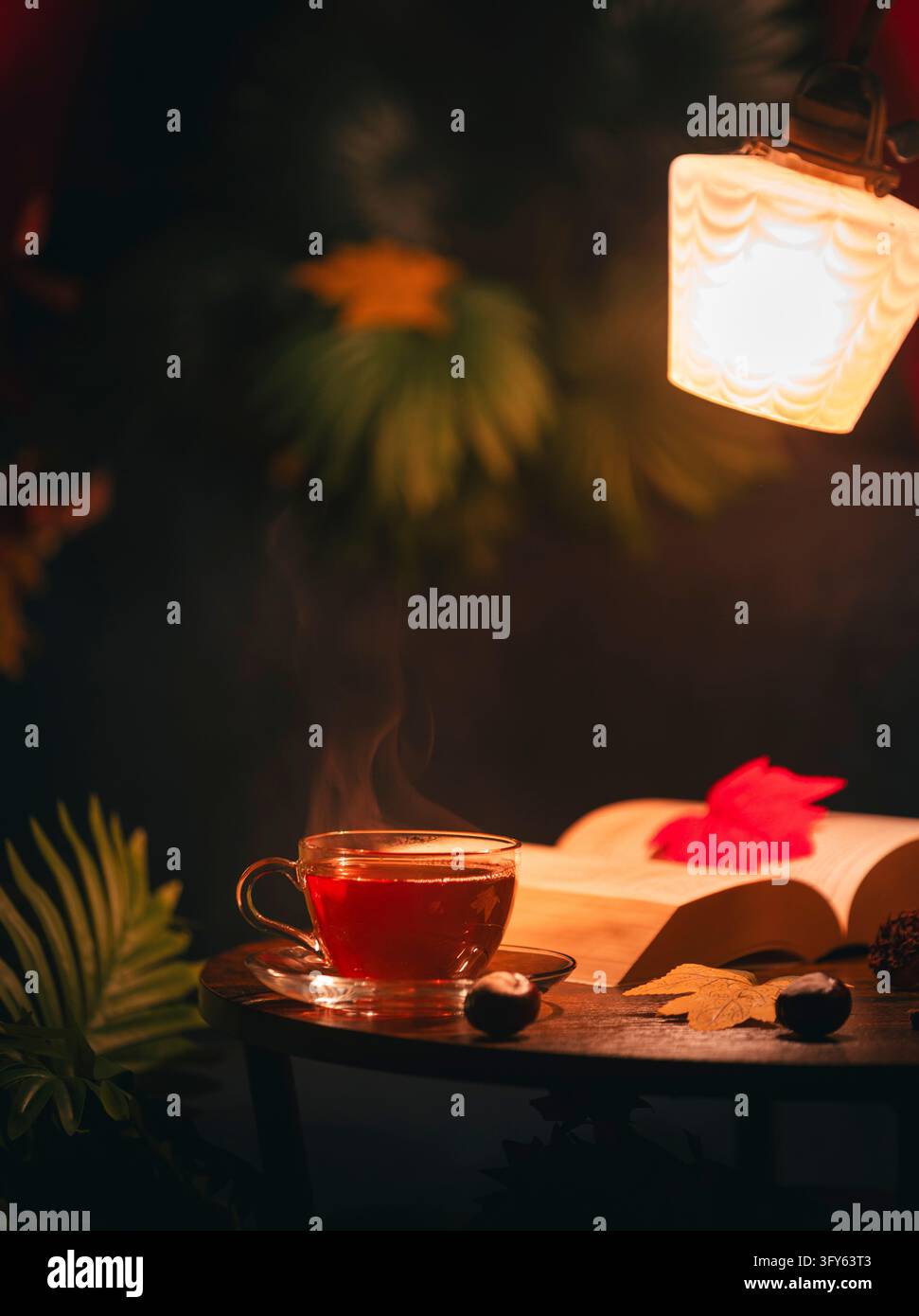 Una tazza di tè e un libro aperto si trovano nella luce calda di una lampada, circondati da foglie autunnali Foto Stock