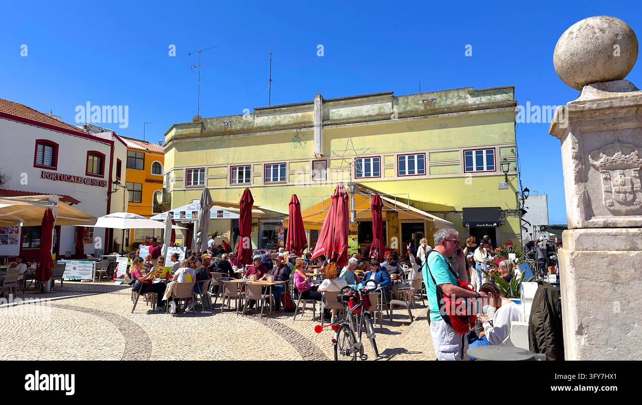 Persone sedute ai tavoli del caffè nella piazza del paese con l'edificio giallo del ristorante Portugalegustus Praça Rainha Dona Leonor Ferragudo, Portimão Portogallo Foto Stock