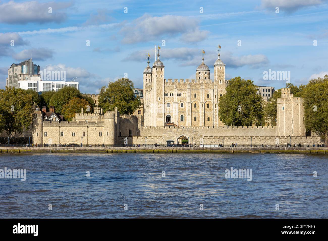 Torri di pietra illuminati dal sole si ergono accanto ad alberi frondosi e edifici moderni, mentre l'acqua ondulata del fiume riempie il primo piano sotto un cielo blu nuvoloso Foto Stock