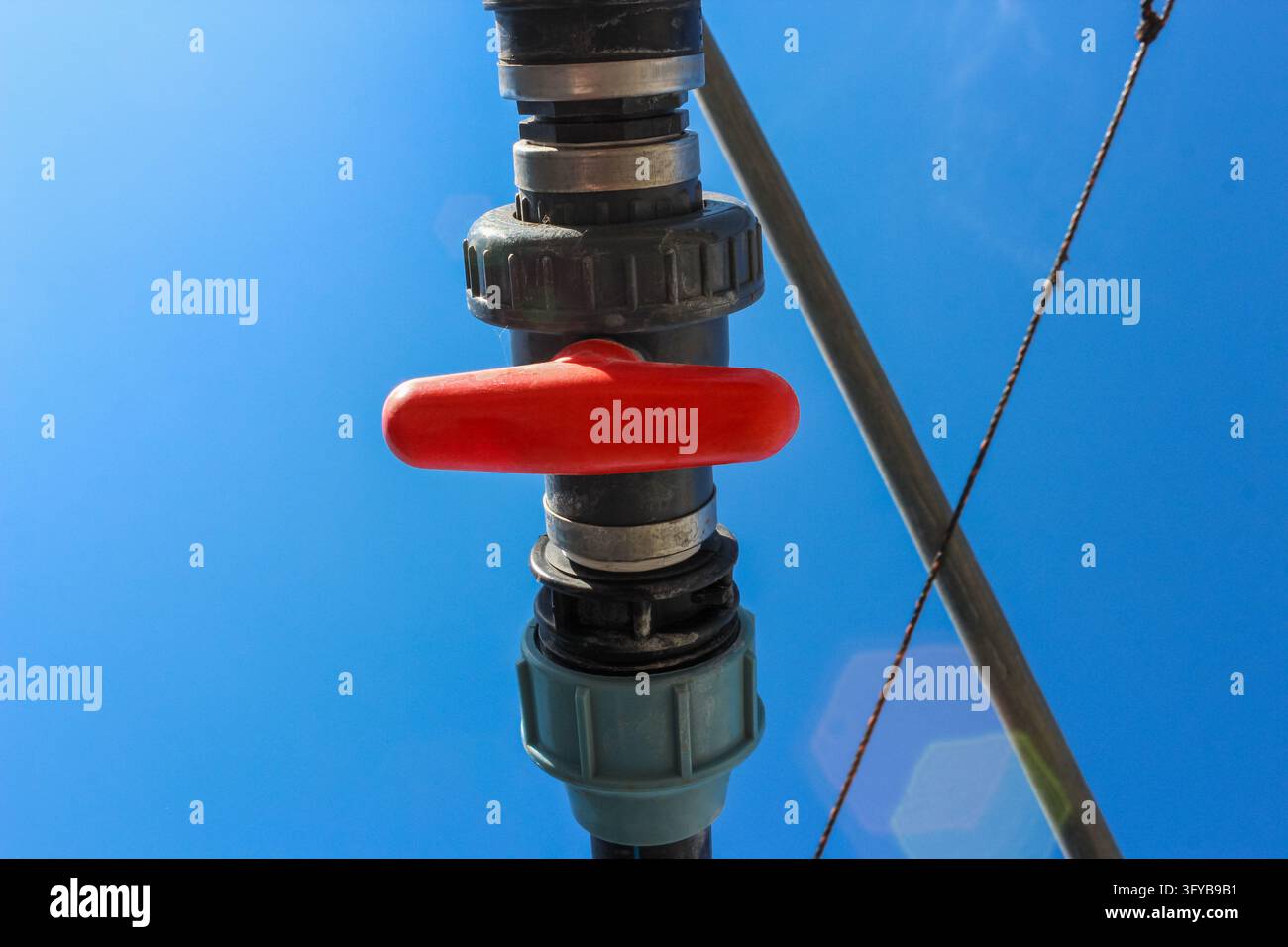 Una valvola a sfera in plastica con maniglia rossa è installata su un tubo di irrigazione nero contro un cielo blu chiaro, attualmente in posizione chiusa. Foto Stock
