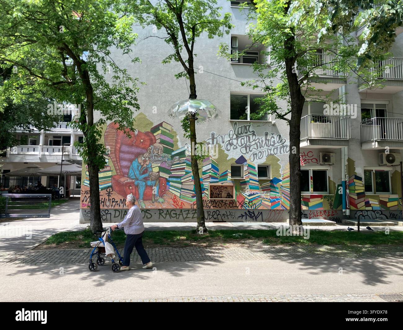 Una persona anziana che usa un deambulatore passa davanti a un murale di street art vibrante sulla facciata di un edificio residenziale a Lubiana, in Slovenia. - Immagine stock catturata con smartphone