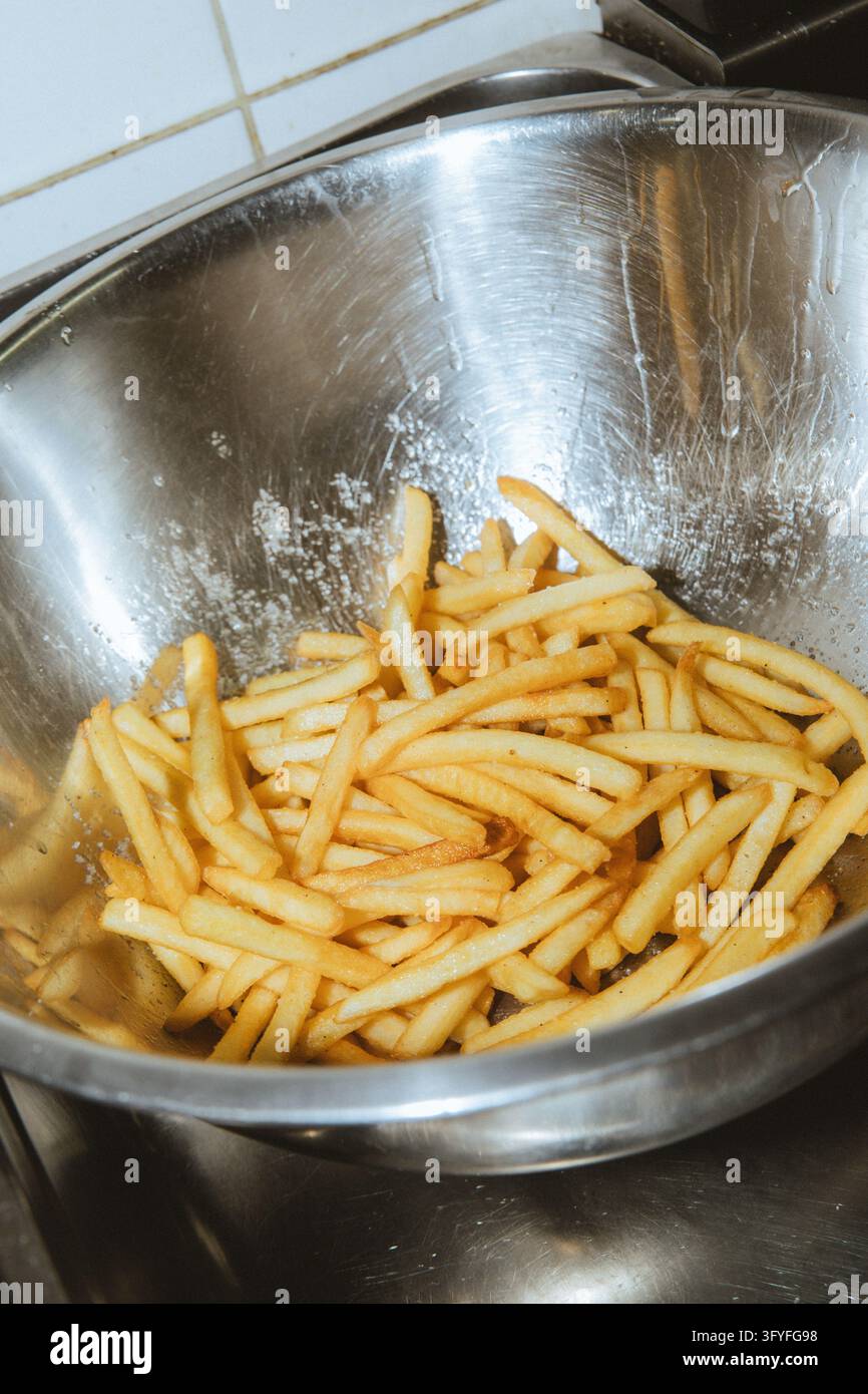 Patatine fritte fresche e croccanti condite in una grande ciotola in acciaio inossidabile all'interno di una cucina di ristorante. Foto Stock