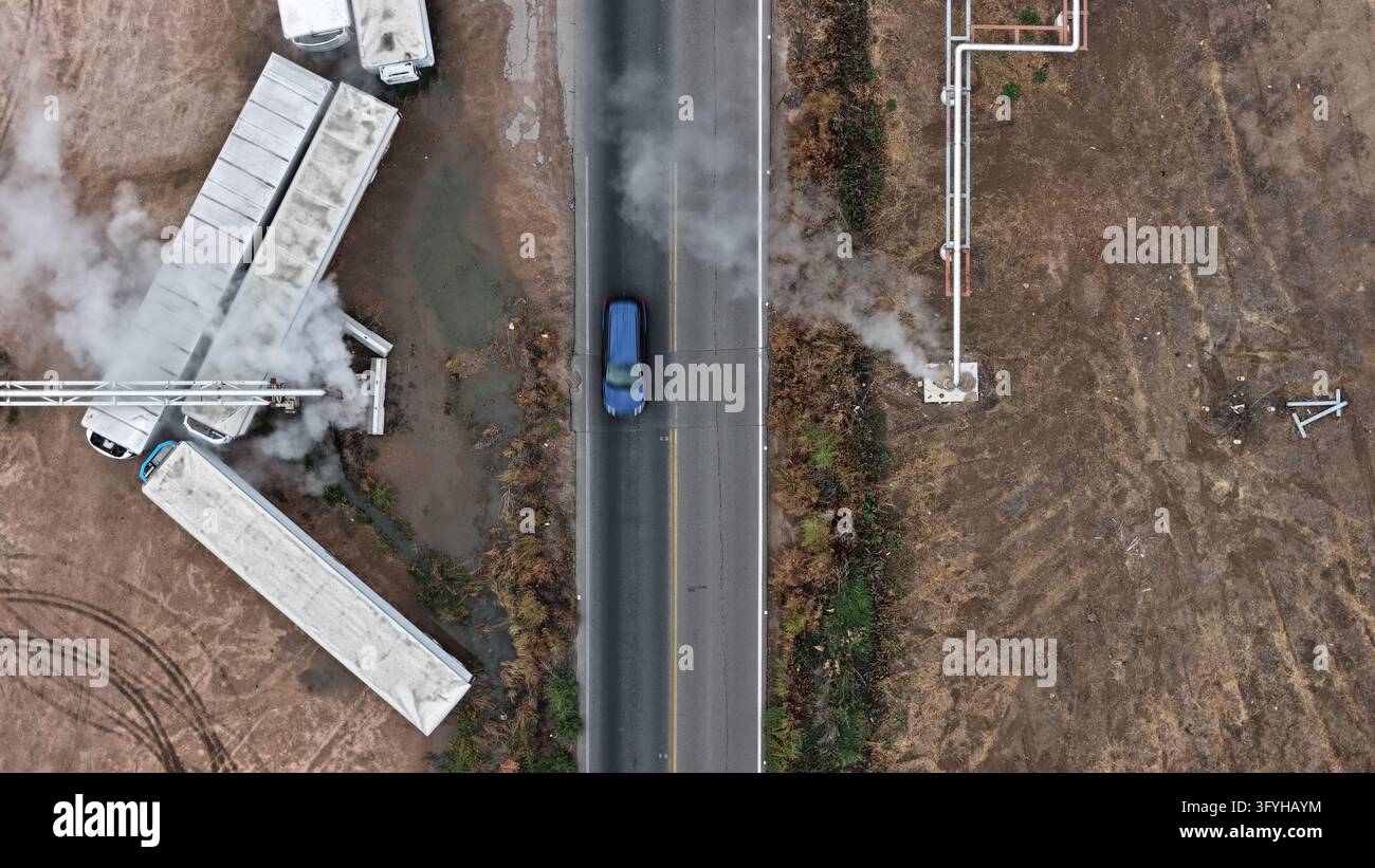 Vista aerea dello stabilimento di lavorazione della carne JBS con tubi industriali che emettono vapore, rimorchi parcheggiati e un'auto blu che percorre la strada adiacente a Gre Foto Stock