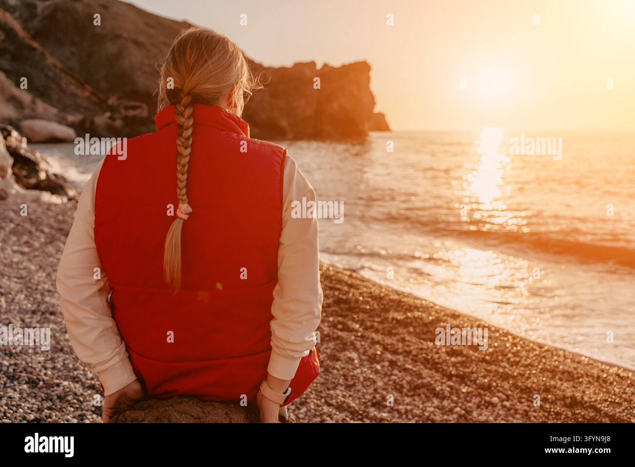 Donna al tramonto sulla spiaggia che guarda il sole dorato sul mare seduta su una riva di ciottoli indossando un giubbotto rosso Foto Stock