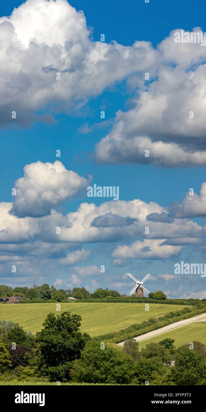 Inghilterra, Wiltshire, Wilton Windmill. Una tradizionale molino a vento si erge oltre terre verdi mentre una strada di campagna sinuosa attraversa campi ondulati sotto un cielo drammatico Foto Stock