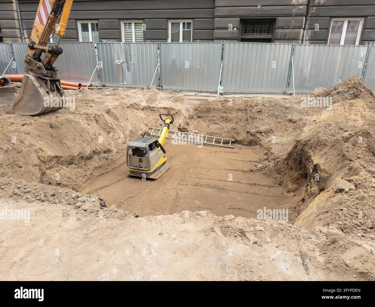 Un compattatore industriale per il suolo poggia su terra livellata all'interno di una trincea in un cantiere edile cittadino affollato, evidenziando i macchinari pesanti utilizzati durante le fondazioni Foto Stock