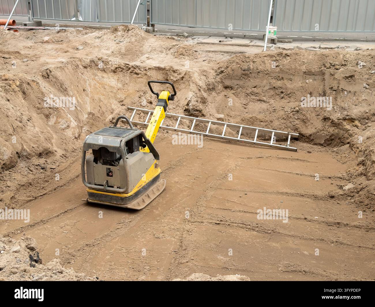 Compattatore a piastra vibrante si trova su terreno instabile all'interno di uno scavo profondo, rappresentando attività di preparazione del terreno e ingegneria industriale per p Foto Stock