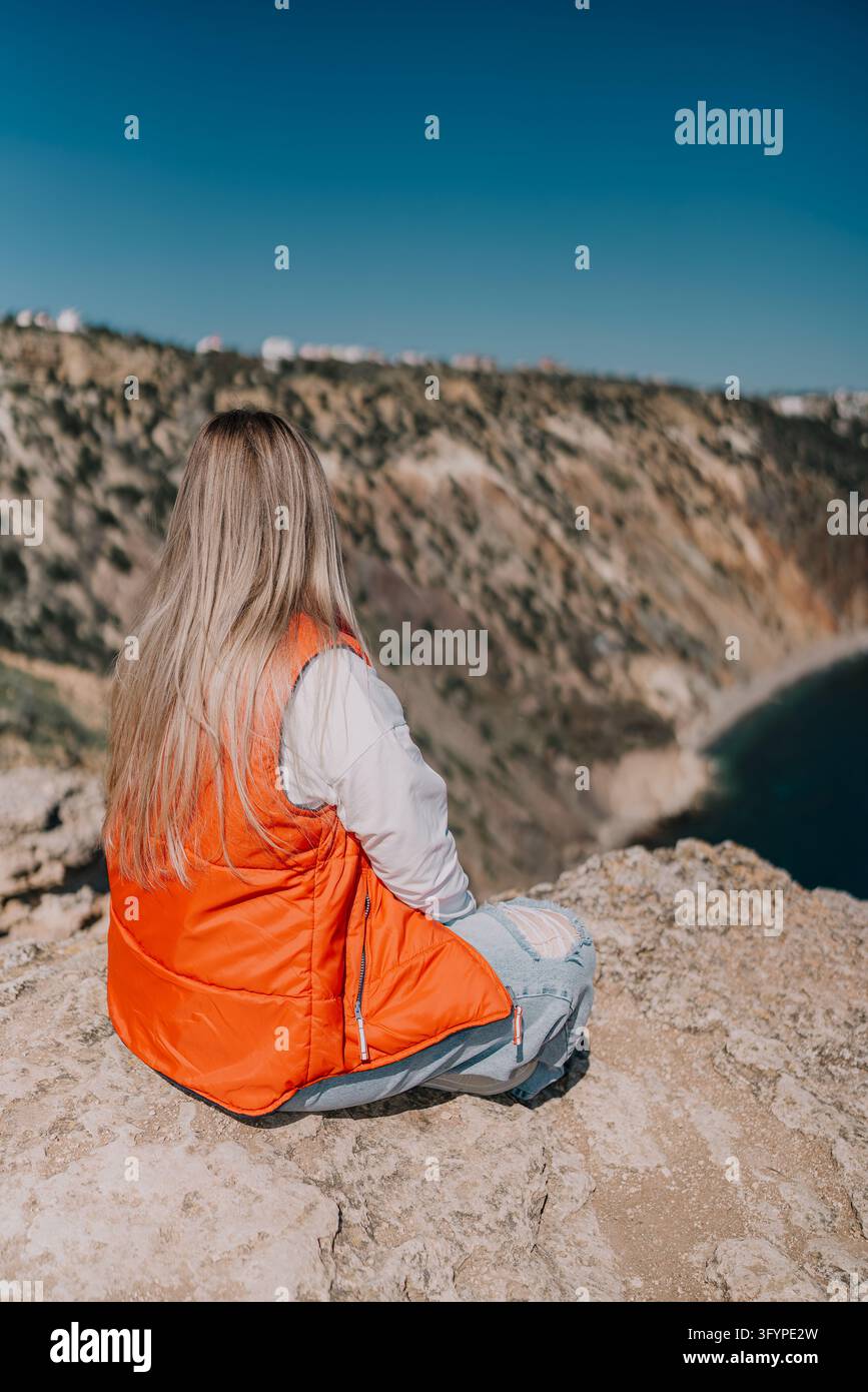 Donna seduta su un bordo roccioso sulla scogliera, indossa un giubbotto arancione e guarda il mare blu panoramico e l'orizzonte Foto Stock