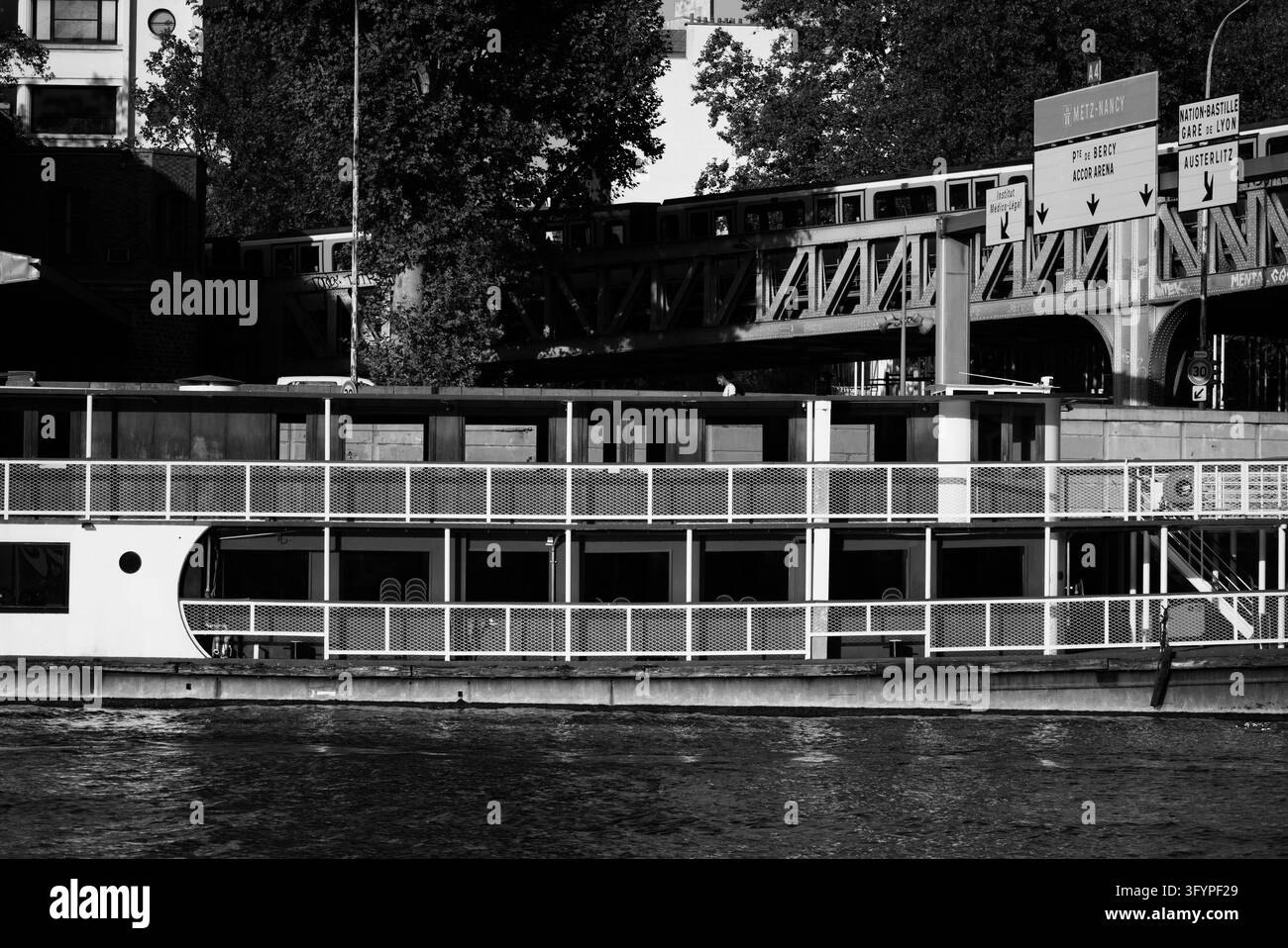 Vista in bianco e nero di una chiatta passeggeri ormeggiata lungo la Senna a Parigi, con binari della metropolitana sopraelevati, segnali stradali per la direzione Austerlitz Foto Stock
