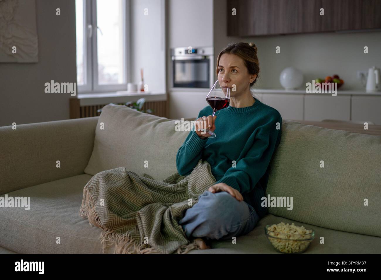 Serata cinema casalinga, intrattenimento in solitaria, momento di relax confortevole, godersi i piaceri semplici dopo una giornata impegnativa. Donna di mezza età interessata che beve vino rosso seduta Foto Stock