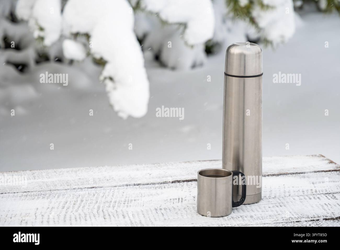 Borraccia sottovuoto in acciaio inossidabile e tazza di metallo su un tavolo di legno coperto di neve con rami sempreverdi ghiacciati sullo sfondo. Composizione invernale all'aperto minimalista Foto Stock