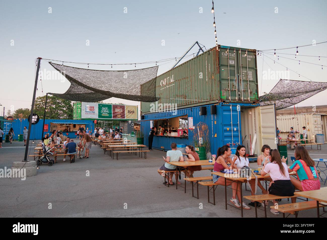 30 agosto 2026, Belgrado, Serbia: Folle che cena in un mercato alimentare in container per navi al crepuscolo, con luminarie e gru portuali sopra. Foto Stock