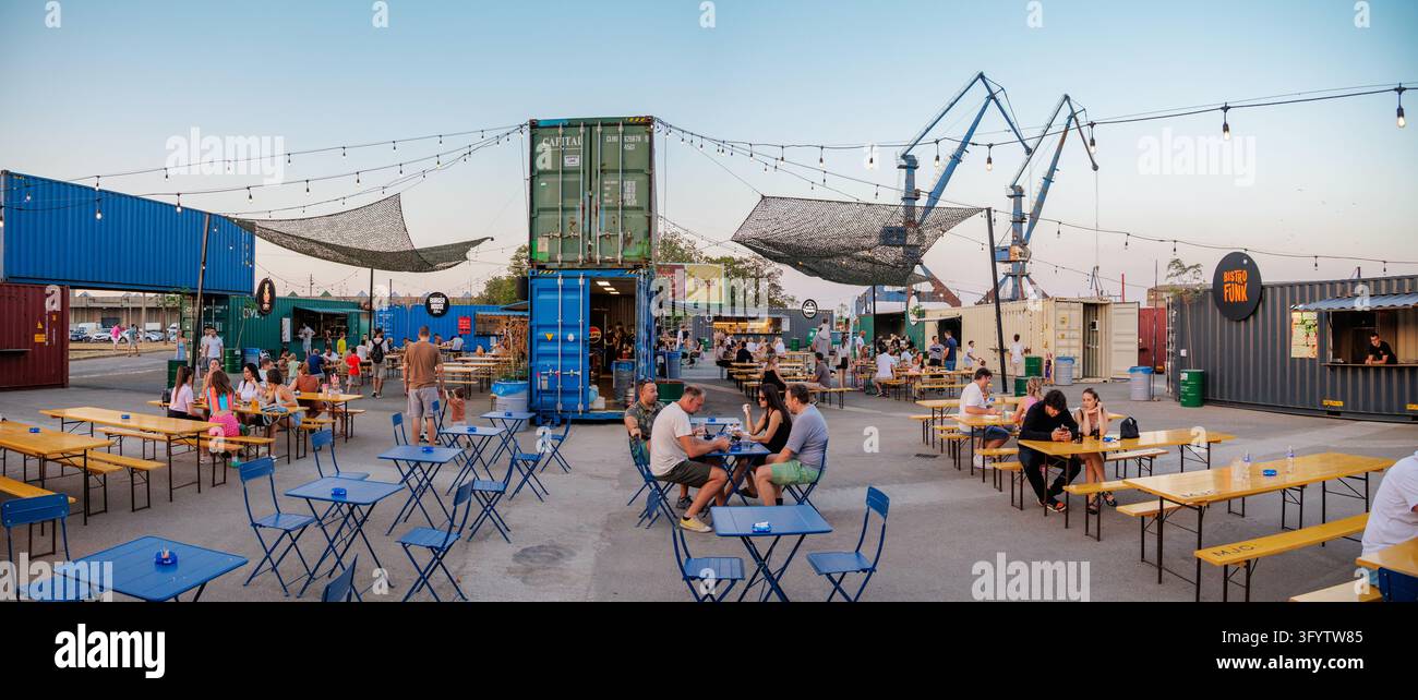 30 agosto 2026, Belgrado, Serbia: Folle che cena in un mercato alimentare in container per navi al crepuscolo, con luminarie e gru portuali sopra. Foto Stock