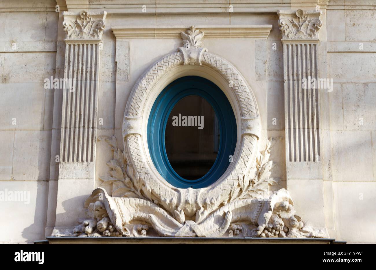 Finestra ovale con cornice blu circondata da una ghirlanda di alloro scolpita e cornucopie tra pilastri scanalati su una facciata in pietra, Parigi, Francia Foto Stock