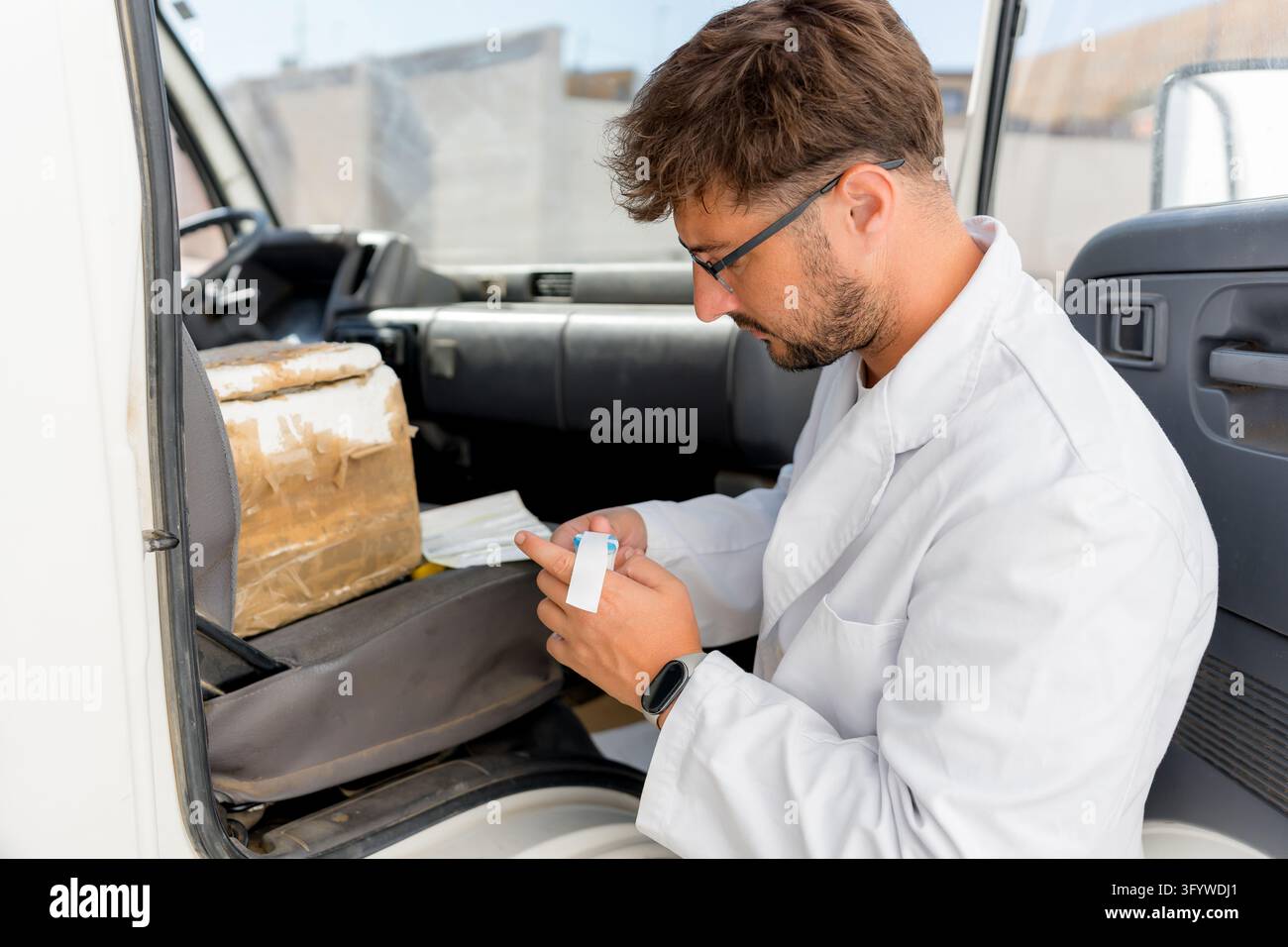 Uomo in camice bianco e occhiali etichetta un piccolo contenitore di latte di pecora accanto a una scatola isolata in un furgone, preparando il campione per l'analisi e il caseificio Foto Stock