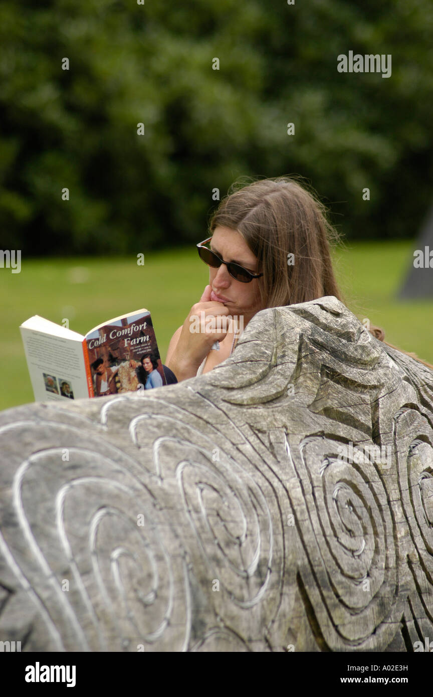 Giovane donna che indossa la lettura sunglassses Cold Comfort Farm libro in brossura seduto scolpito sul banco di legno Machynlleth powys GALLES Foto Stock