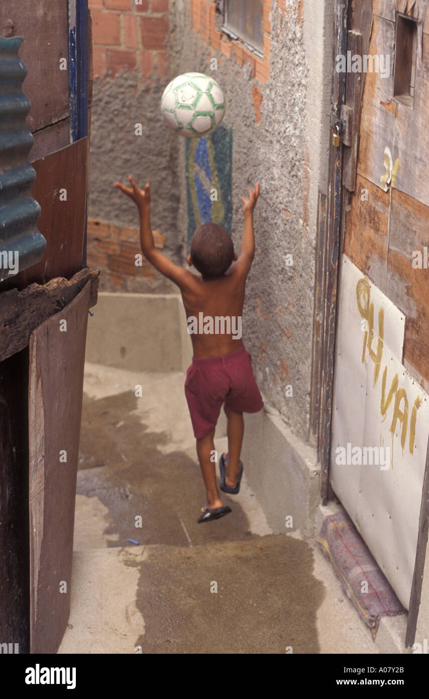 Vita quotidiana a Rio de Janeiro favela, 10 anni di età gioca con la palla di calcio a Morro da Mangueira (Mangueira slum) - fogna aperta in vicolo stretto. Foto Stock