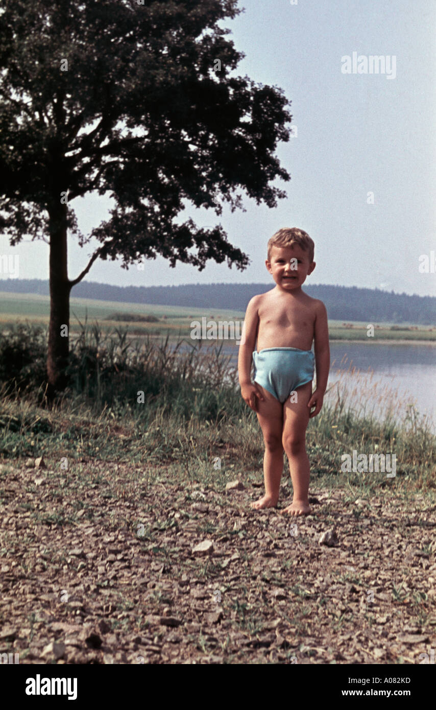 'Pronto per niente". Un ragazzo in piedi sulla riva di un lago con la sua swimpants rivolta verso la telecamera Foto Stock