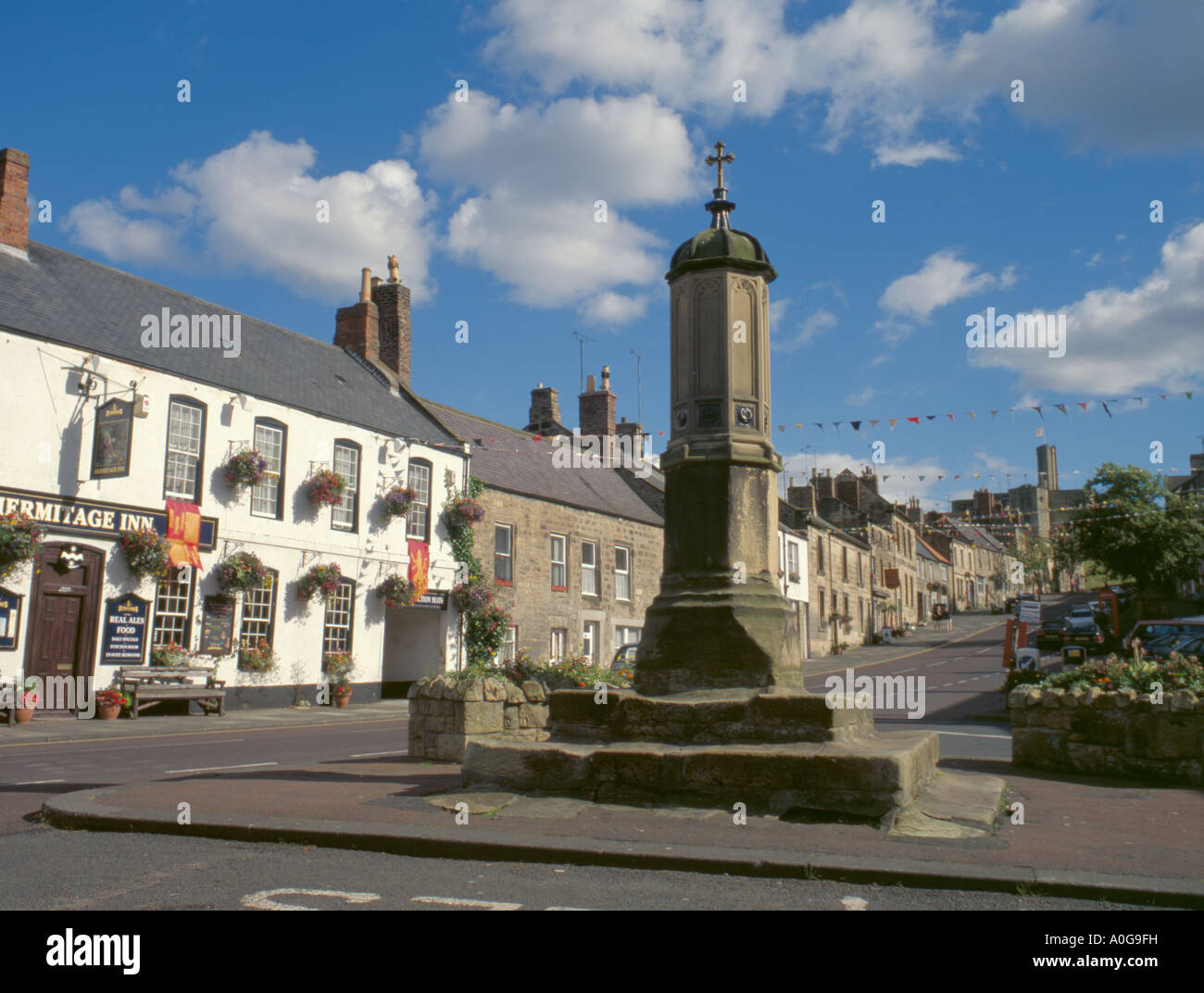 Mercato di arenaria croce e borgo medievale di warkworth con il castello di warkworth oltre, Northumberland, Inghilterra, Regno Unito. Foto Stock