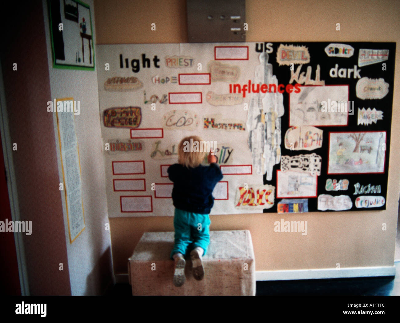 Piccolo ragazzo biondo guardando un collage parete in una sala della scuola Foto Stock
