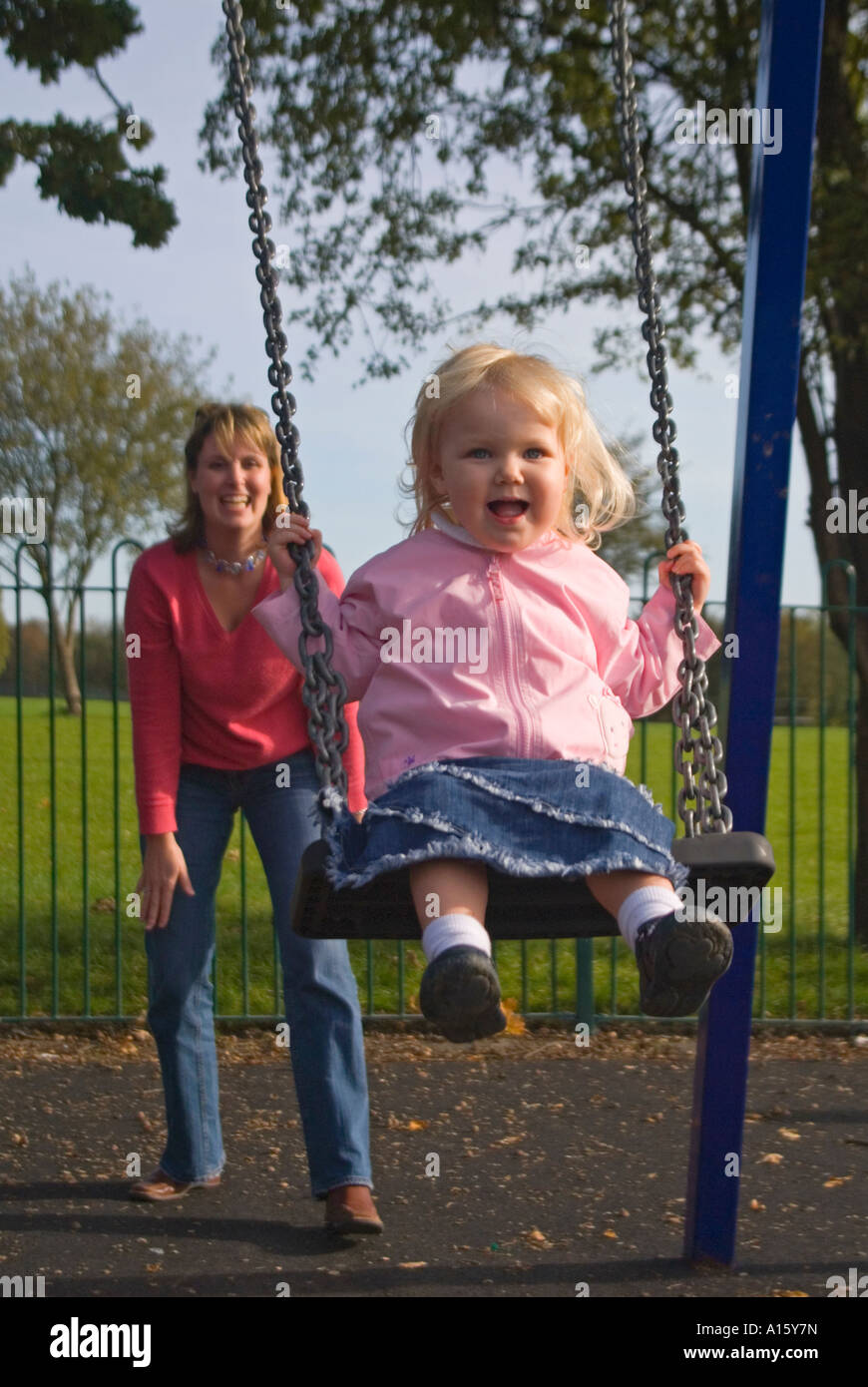 Ritratto verticale di una giovane madre caucasica spingendo la sua bionda bambina su altalena nel parco. Foto Stock