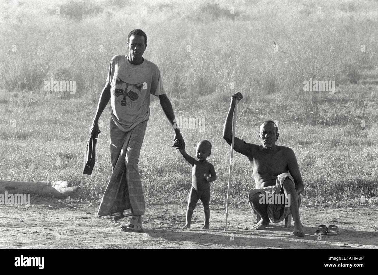 B/N di due uomini ORMA e di un ragazzino, il più giovane con una radio portatile e l'anziano che squattava con un bastone, alla luce del tardo pomeriggio. Wajir, Kenya Foto Stock
