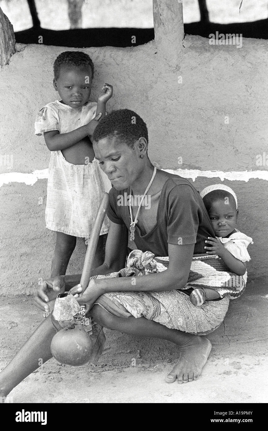 B/N di una donna che fuma un tradizionale tubo d'acqua mentre aspetta con i suoi bambini fuori di una clinica di salute rurale. Vicino Binga, Zimbabwe Foto Stock