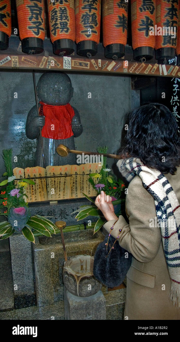 Donna Giapponese in cappotto e sciarpa versando acqua su scolpiti Jizo statua a un tempio nel centro di Kyoto in Giappone Foto Stock
