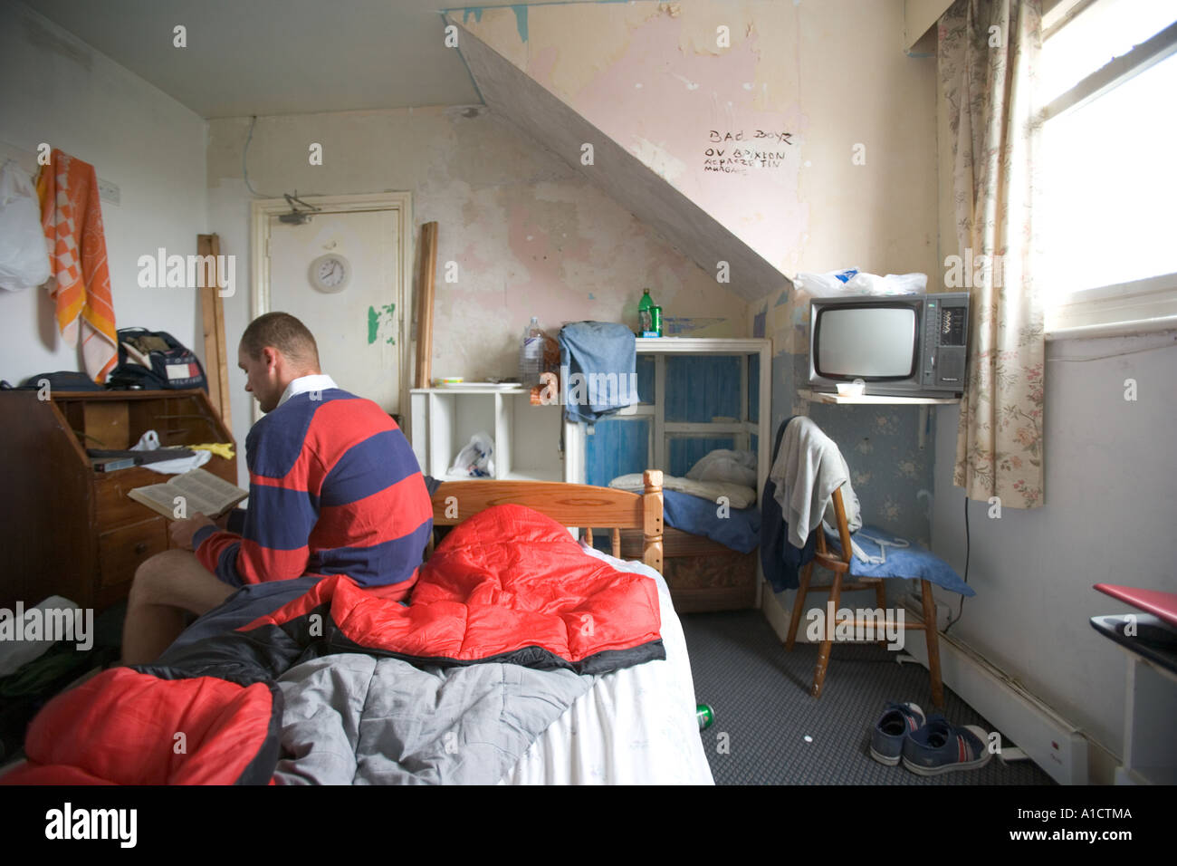 Giovane uomo seduto nel letto a leggere un libro in una piccola camera disordinati Foto Stock