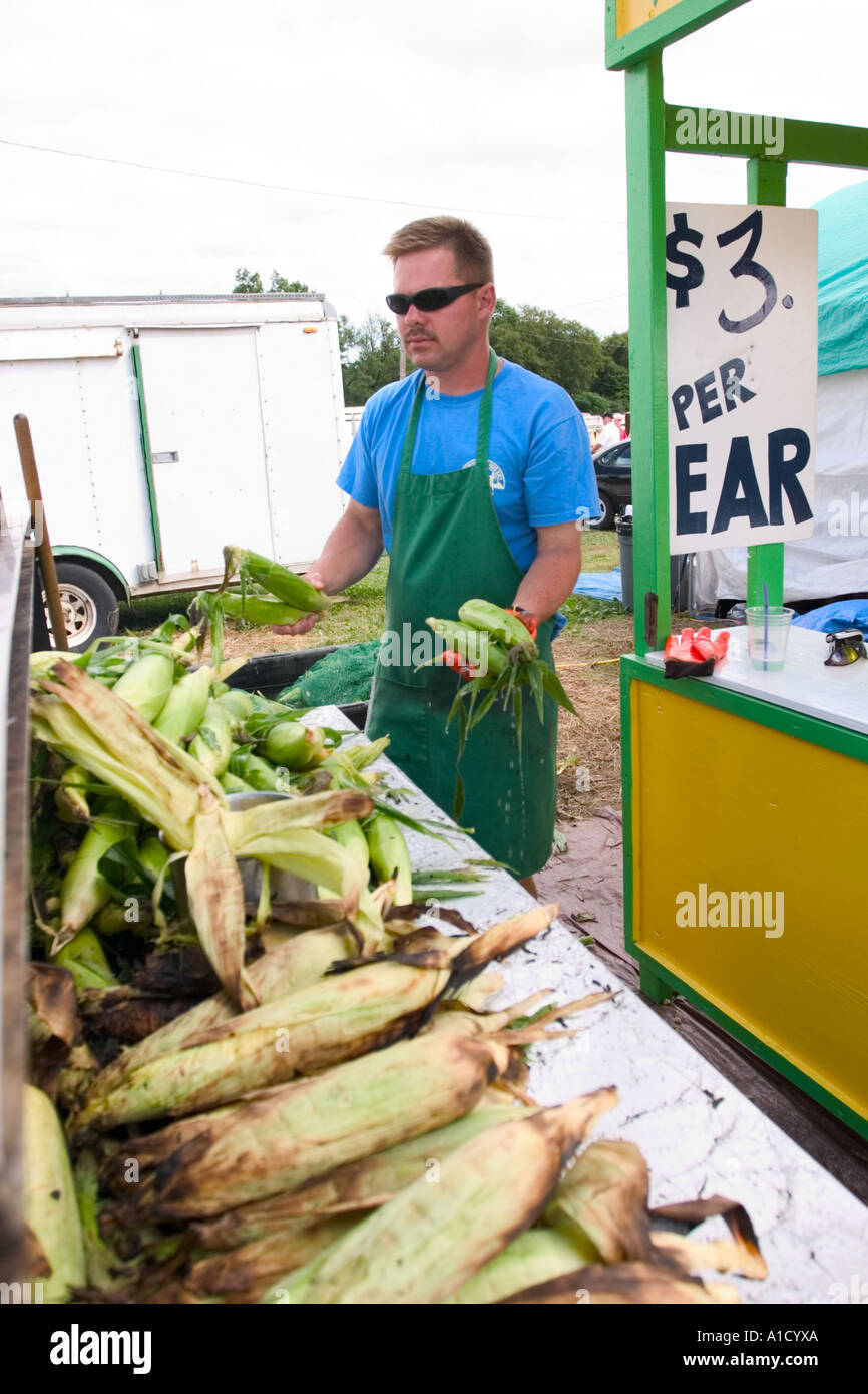 Fornitore maschio ad una fiera in Connecticut USA tenendo un orecchio di mais accanto a un grill dove egli è la tostatura altre spighe di grano. Foto Stock