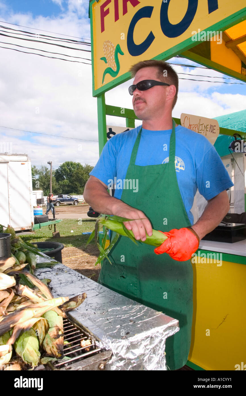Fornitore maschio ad una fiera in Connecticut USA tenendo un orecchio di mais accanto a un grill dove egli è la tostatura altre spighe di grano. Foto Stock