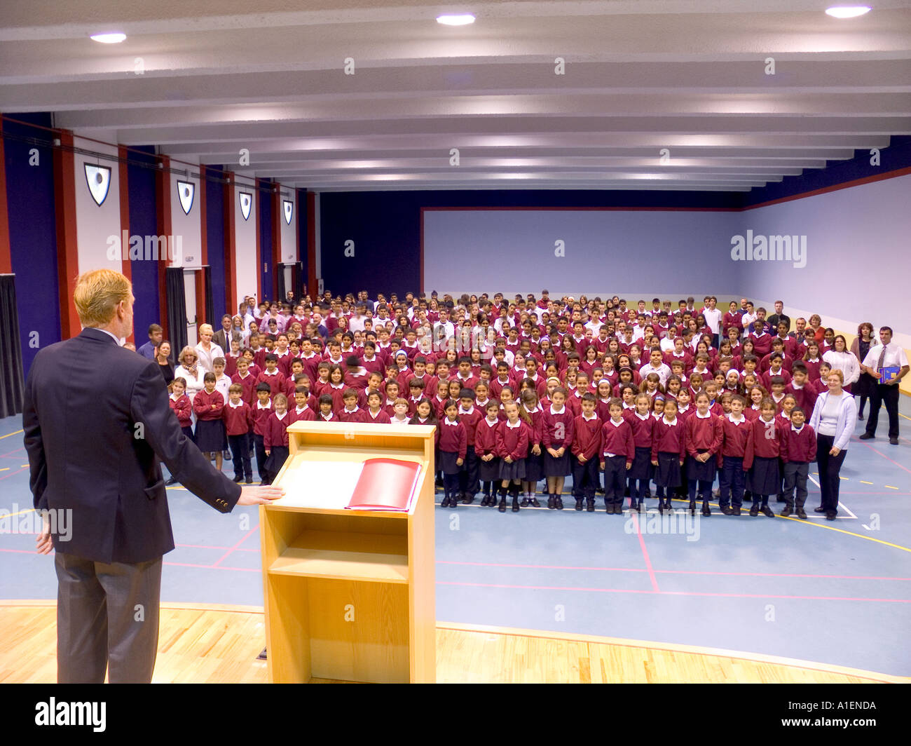Assemblea di istituto capo maestro maestro a leggio prende gli studenti e il personale complessivo nella scuola moderna hall Foto Stock
