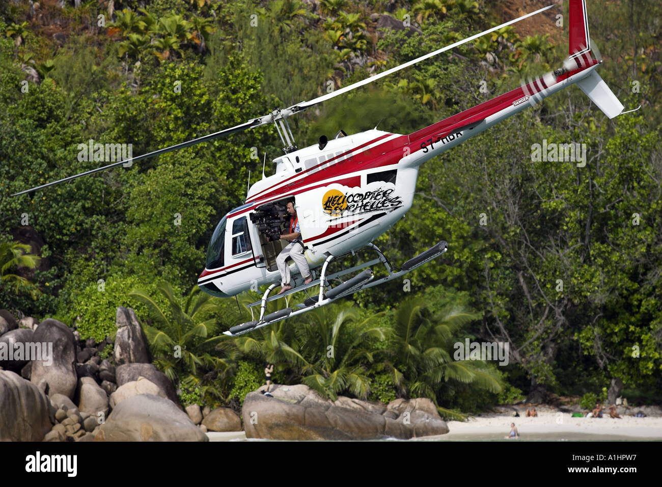Un elicottero che vola basso e vicino alla riva con un cameraman / fotografo seduto alla porta aperta Foto Stock