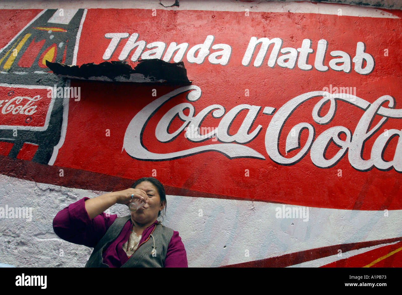 Una donna tibetana si siede di fronte a una grande coca cola pubblicità dipinta su un muro in Bodhgaya, in Bihar in India Foto Stock