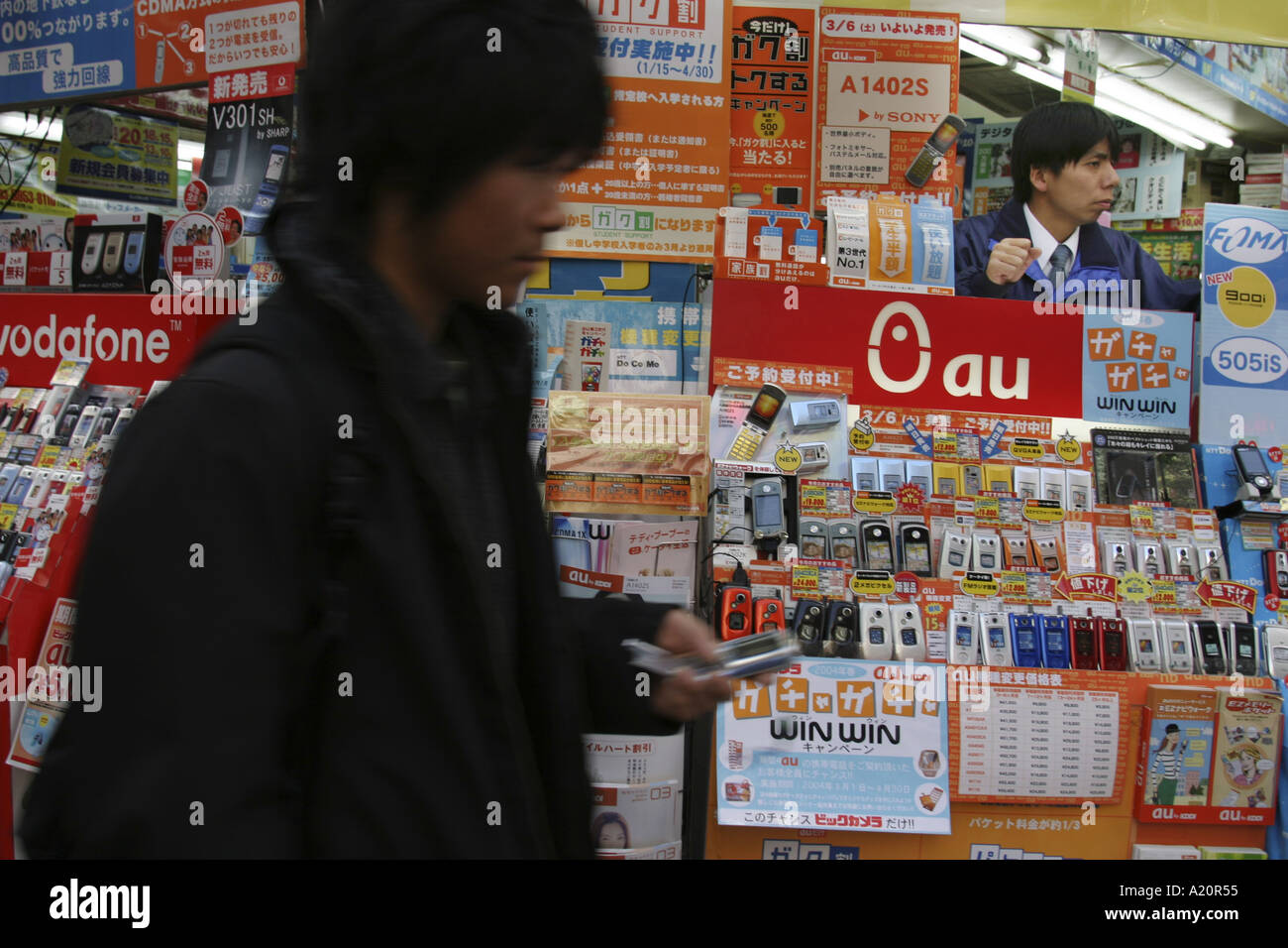 Giovane uomo texting sul suo cellulare passeggiate passato i telefoni mobili per la vendita in un negozio di elettronica, Tokyo, Giappone. Foto Stock