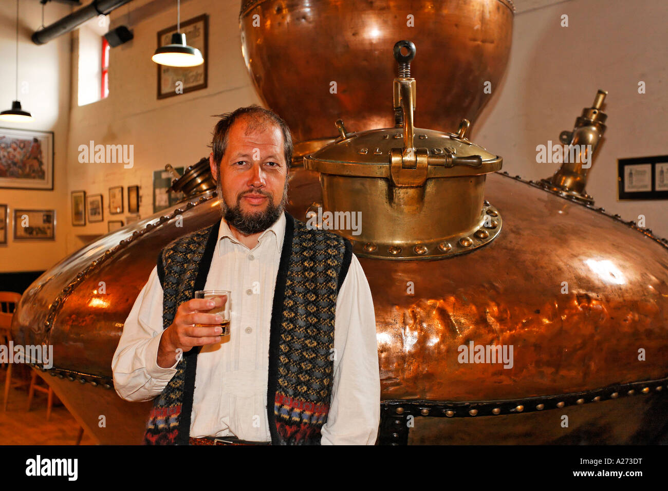 Un uomo gode di un buon bicchiere di whiskey dietro un vecchio Pot still (un gigante bollitore di rame) utilizzato per la distillazione in bushmills Foto Stock