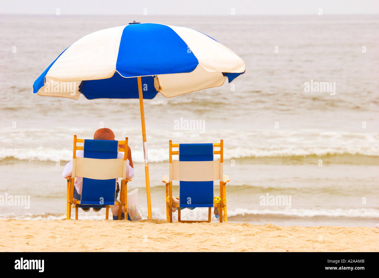 Un uomo solitario si siede su una sedia spiaggia sotto un blu e bianco ombrello, in attesa che il sole a brillare. Foto Stock