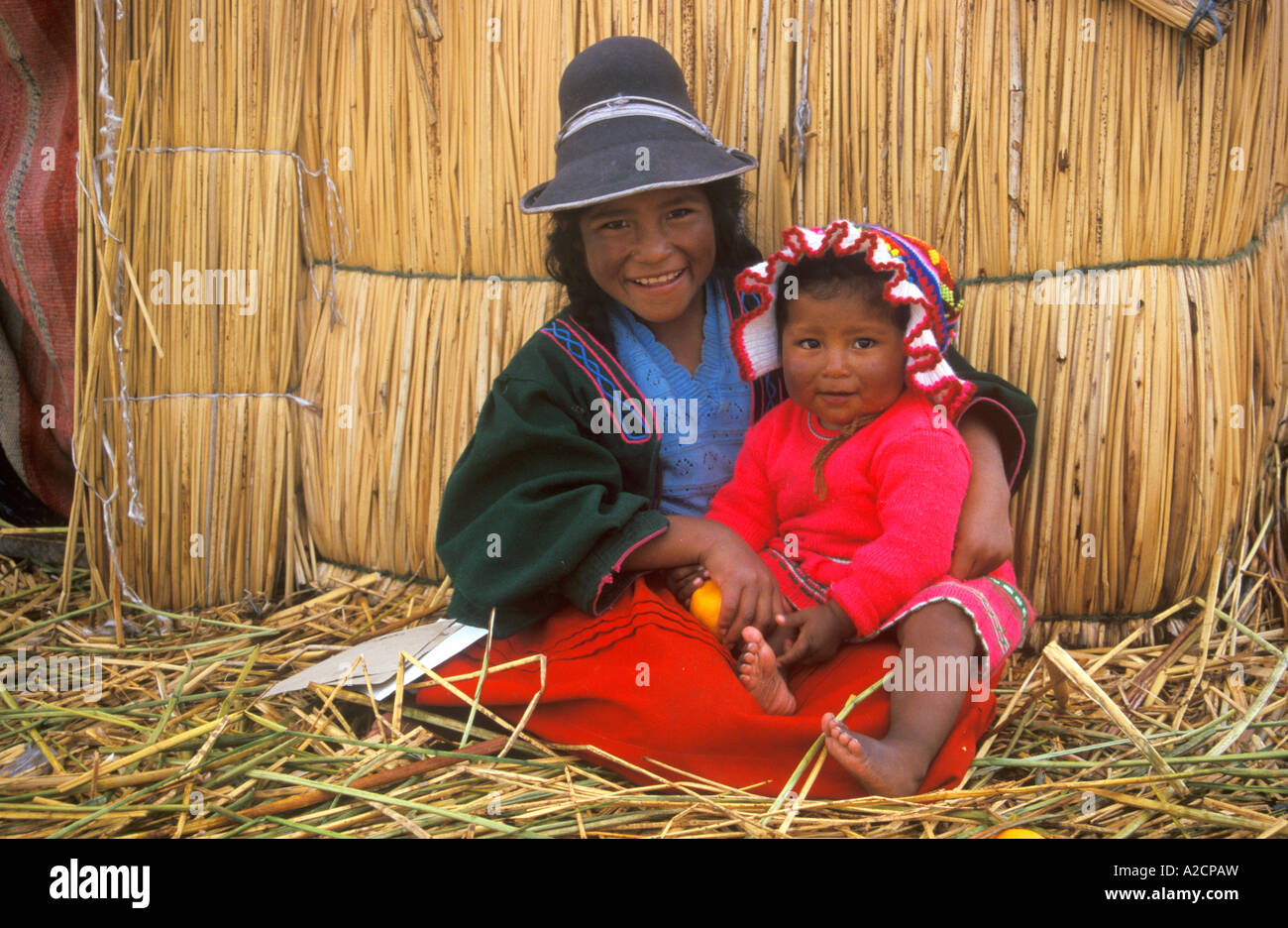 Tradizionalmente vestiti bambini seduti accanto a una capanna di paglia su un Uro Isola una delle numerose isole galleggianti nel Lago Titicaca Foto Stock