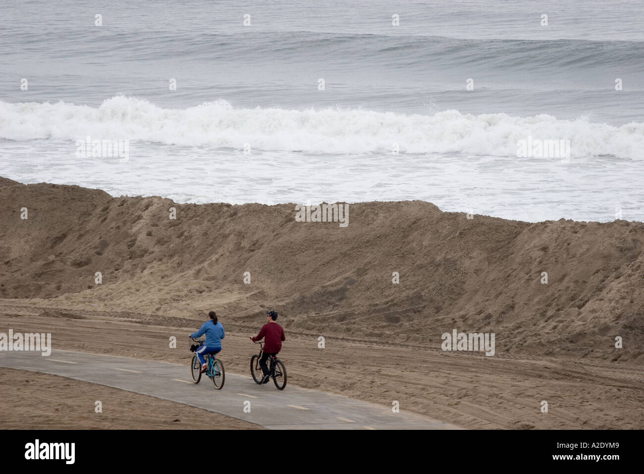 Due piloti di bicicletta pedala sul percorso vicino alla sabbia temporanea berm che protegge le spiagge da sopra la media di onde alte Foto Stock