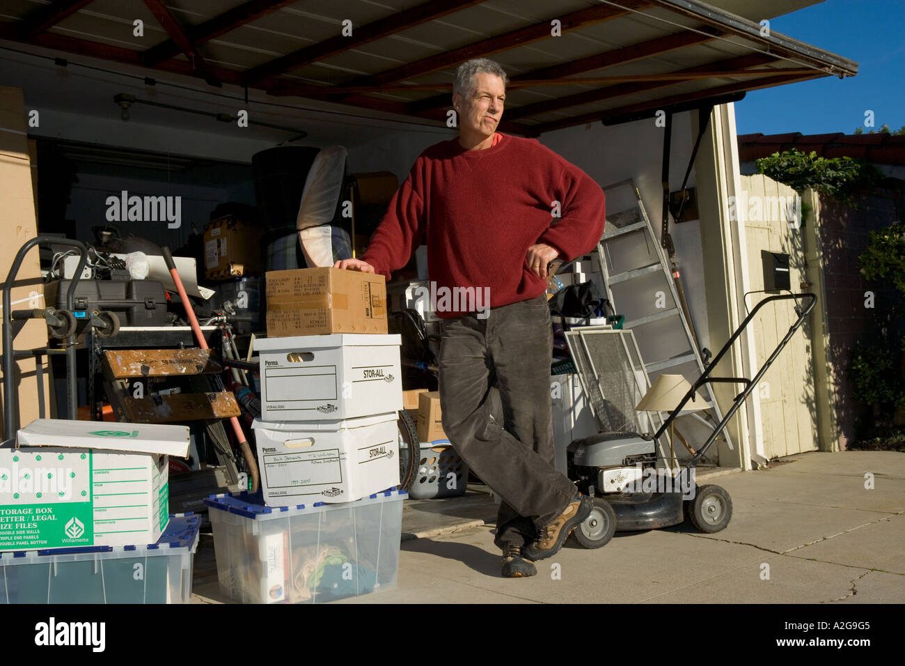 Uomo di mezza età si erge e medita cosa fare con pile di averi nel suo garage sovraffollate Foto Stock